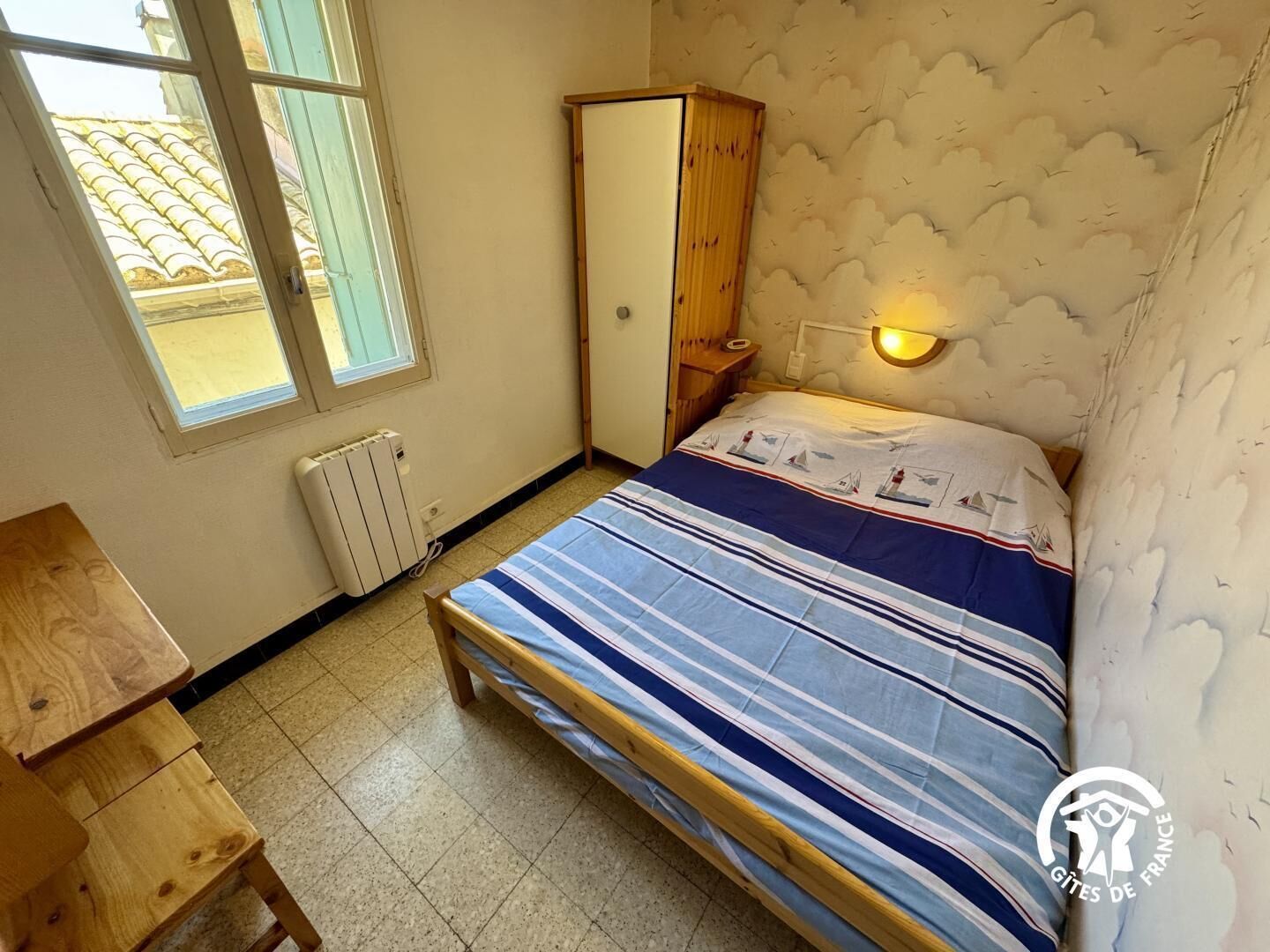A bedroom with a bed, a wooden wardrobe, a nightstand, a wall-mounted light, a radiator, and a window with a view of a tiled roof.