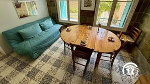 A cozy room with a teal sofa, a wooden dining table with chairs, and a large window with a view of greenery. - Nature Sauvage Méditerranée (BAGES)