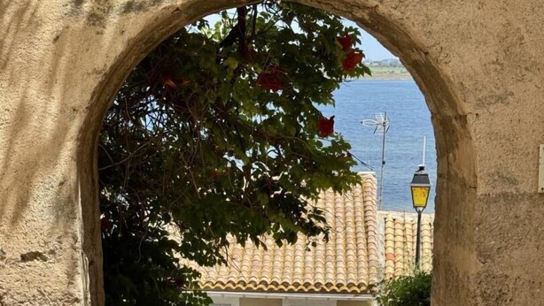 A stone archway leading to a building with a tiled roof, a small garden with plants, and a signboard. - Nature Sauvage Méditerranée (BAGES)
