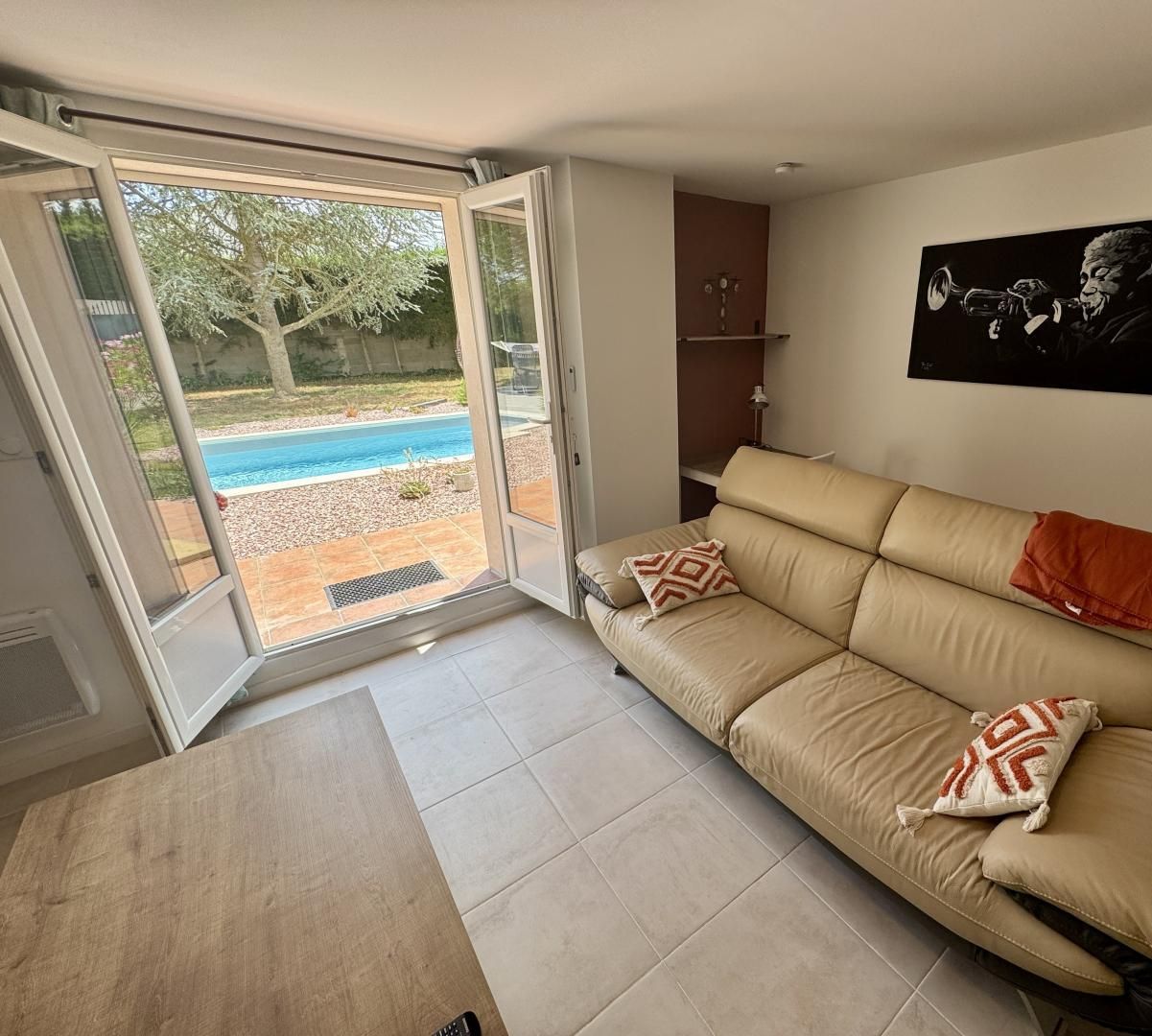 A modern living room with a beige sofa, two decorative pillows, a glass door leading to a pool, and a framed artwork on the wall.