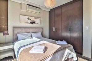 A bedroom with a bed, bedside table, lamp, air conditioning unit, wardrobe, and artwork on the wall.