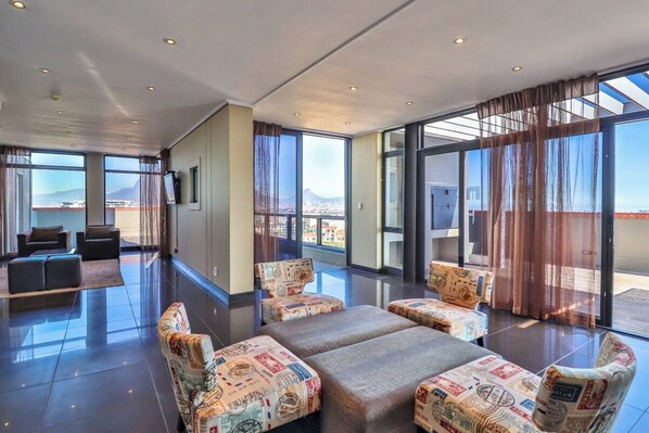 A spacious room with a large bed, two armchairs, and a sofa. There are large windows offering a view of the city and mountains.