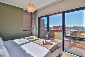 A hotel room with a large bed, a view of the city through floor-to-ceiling windows, and a balcony with a view of the rooftops.