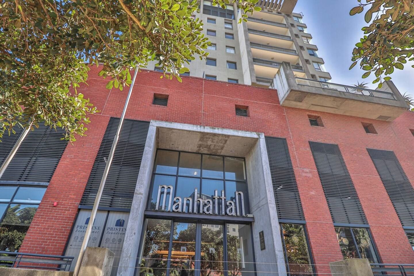 Un immeuble dont l’entrée est marquée par une enseigne bien visible indiquant « Manhattan », caractérisé par un style architectural moderne, de grandes fenêtres et une façade en briques rouges.