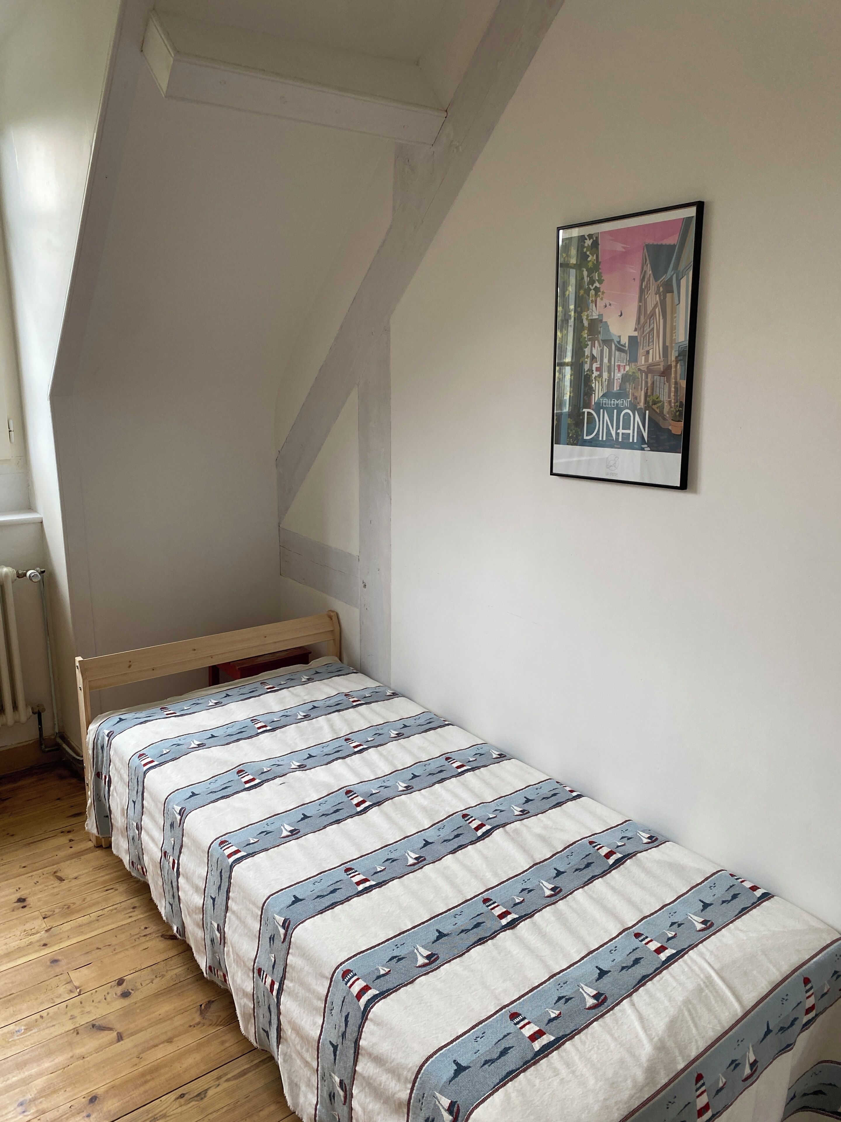 A small, single-bed room with a sloped ceiling and a radiator on the left. A framed picture of a cityscape hangs on the wall.