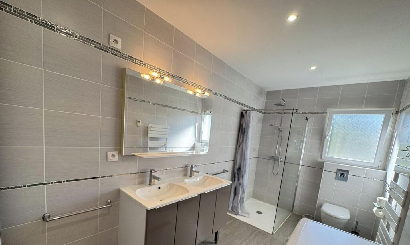 A modern bathroom with a double sink vanity, a glass-enclosed shower, and a window providing natural light.