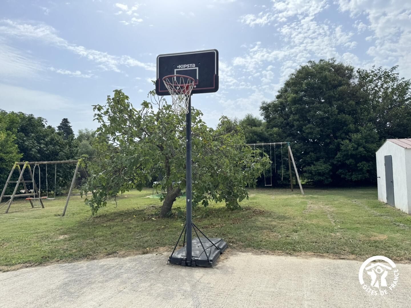 Canasta de baloncesto con logotipo 'KIPSTA', un conjunto de columpios y un cobertizo pequeño en un área verde.
