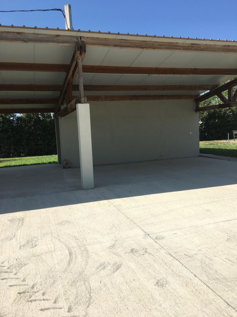 Un espacio al aire libre techado con suelo de hormigón, vigas de madera y un pilar blanco.
