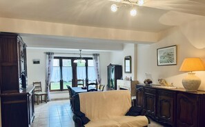 Ein großzügiges Wohnzimmer mit einer Couch, einem Esstisch und einem Kamin. Ein großes Fenster mit Vorhängen, einer Chandelieber und einer Lampe auf einem Beistelltisch.
