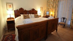 Een traditioneel houten bed met hemelbed, witte lakens, twee nachtkastjes met lampen, een stoel en bloemrijke gordijnen.