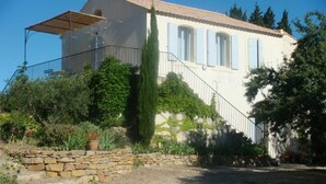 Une maison à deux étages avec un escalier en pierre menant à un balcon, entourée de verdure et sous un ciel dégagé.