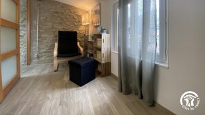 A room with a stone wall, a black chair, a small ottoman, a lamp, and a window with curtains. - Nicou's house - CLARA-VILLERACH (CLARA-VILLERACH)