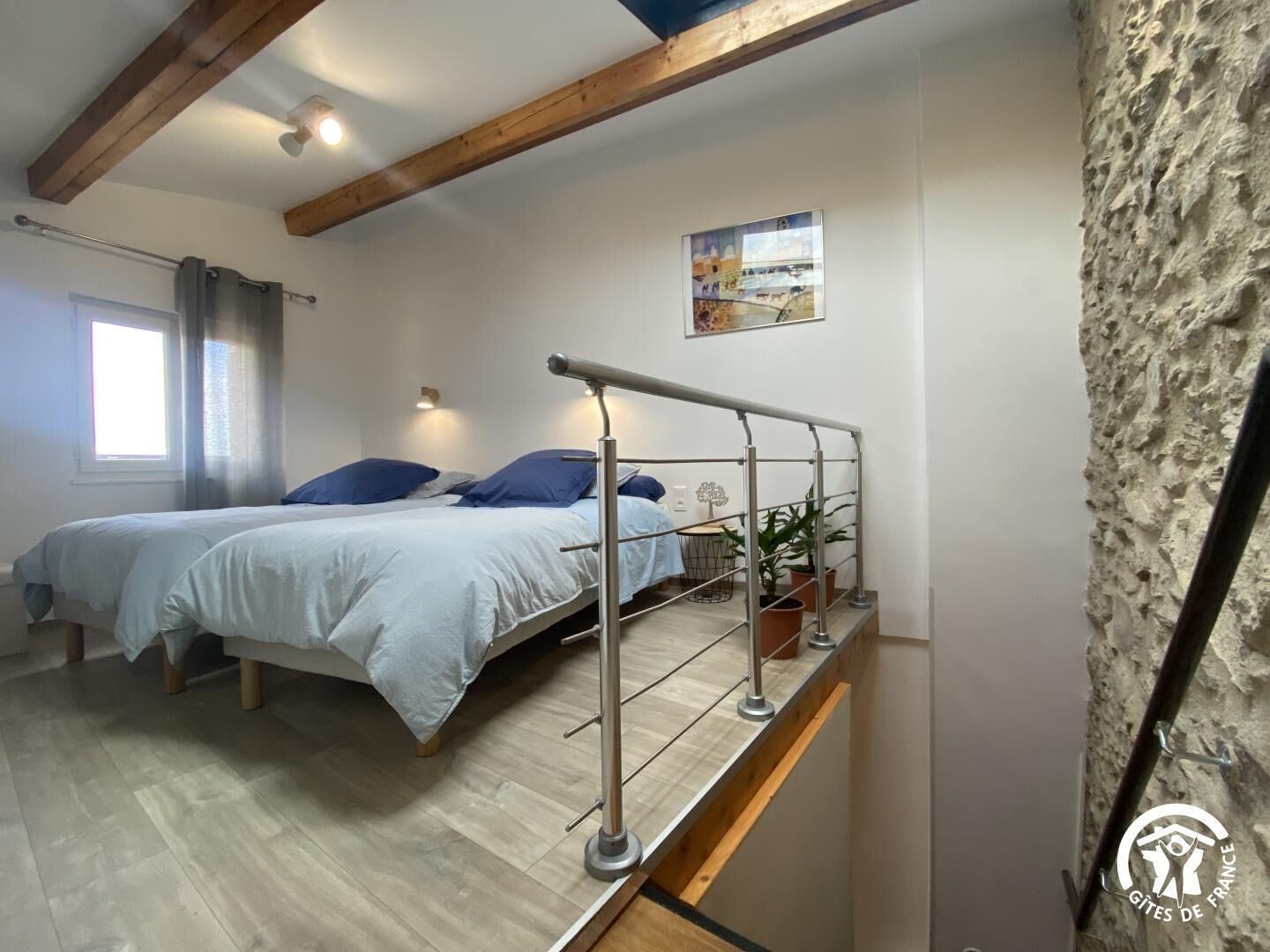 A bedroom with two beds, a wooden ceiling, a window with curtains, a wall-mounted light, and a small plant on a shelf.