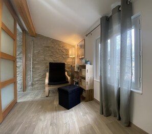 A room with a stone wall, a black chair, a small ottoman, a lamp, and a window with curtains.