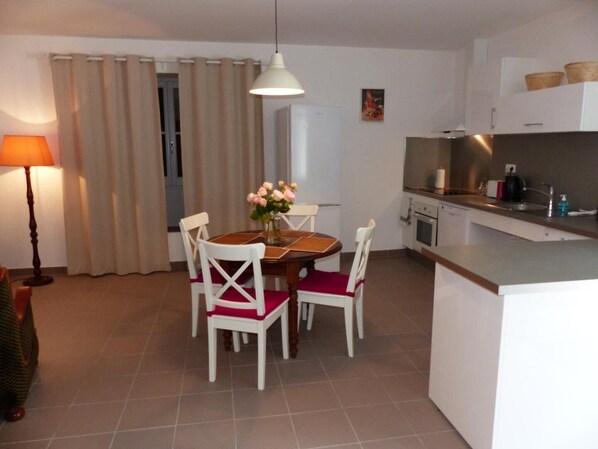 A compact kitchen with a dining area, featuring a table with chairs, a hanging light, and a refrigerator.