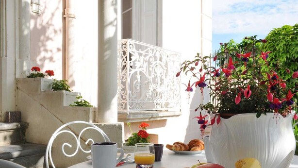 Ein Tisch mit einer weißen Tasse, einem Glas Orangensaft, einem Teller mit Croissants und einer großen weißen Vase mit Blumen.