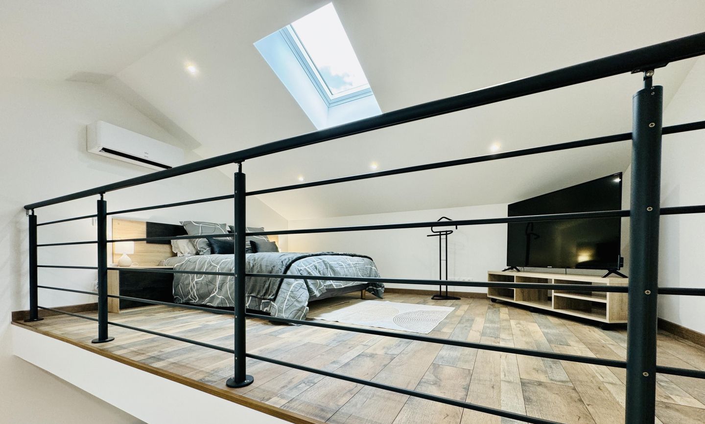 A modern loft bedroom with a skylight, a bed with a gray patterned duvet, a TV, and a wooden floor.
