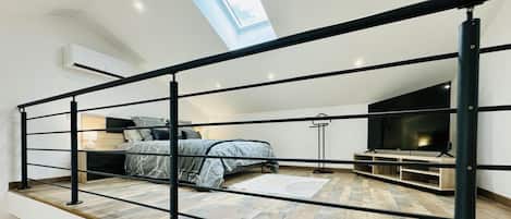 A modern loft bedroom with a skylight, a bed with a gray patterned duvet, a TV, and a wooden floor.