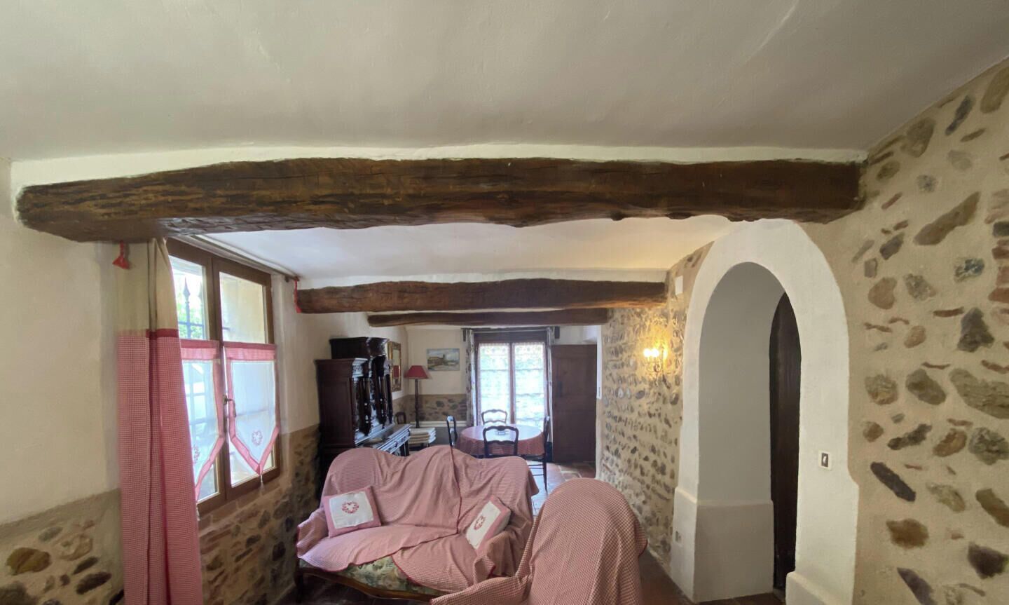 Ein gemütliches Zimmer mit Steinwand, einem Holzbalken an der Decke, einem Bett mit rosa kariertem Überwurf und einem kleinen Tisch mit rotem Stuhl.