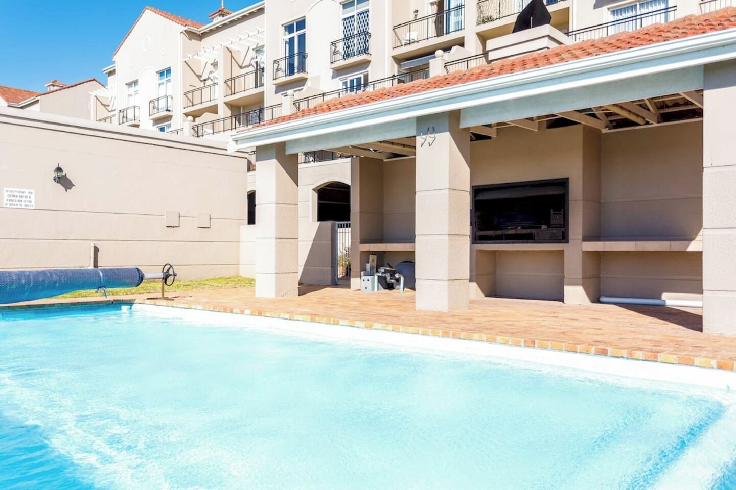 A swimming pool with a covered patio area, a barbecue grill, and a building with balconies.