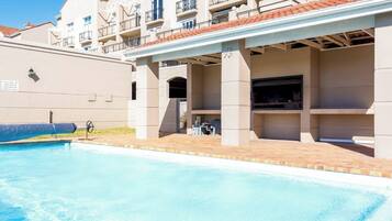 A swimming pool with a covered patio area, a barbecue grill, and a building with balconies.