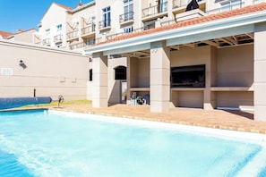 Une piscine avec un patio couvert, un barbecue et un bâtiment doté de balcons.