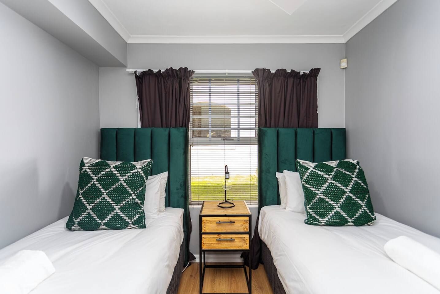 A room with two beds, each with a white mattress and a green patterned pillow, flanked by dark green curtains and a window with blinds.