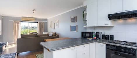 A modern kitchen with white cabinets, a granite countertop, a built-in oven, and a dining area with a sofa and a view of greenery.