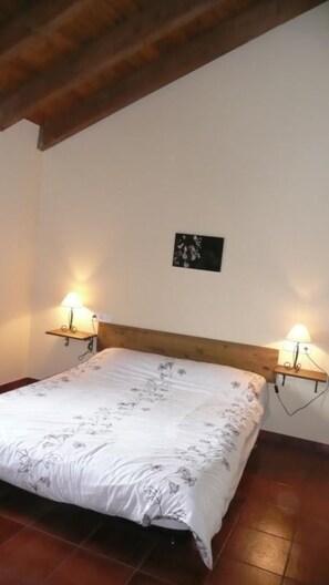 A bedroom with a wooden headboard, a bed with floral bedding, two wall-mounted lamps, and a small picture on the wall.