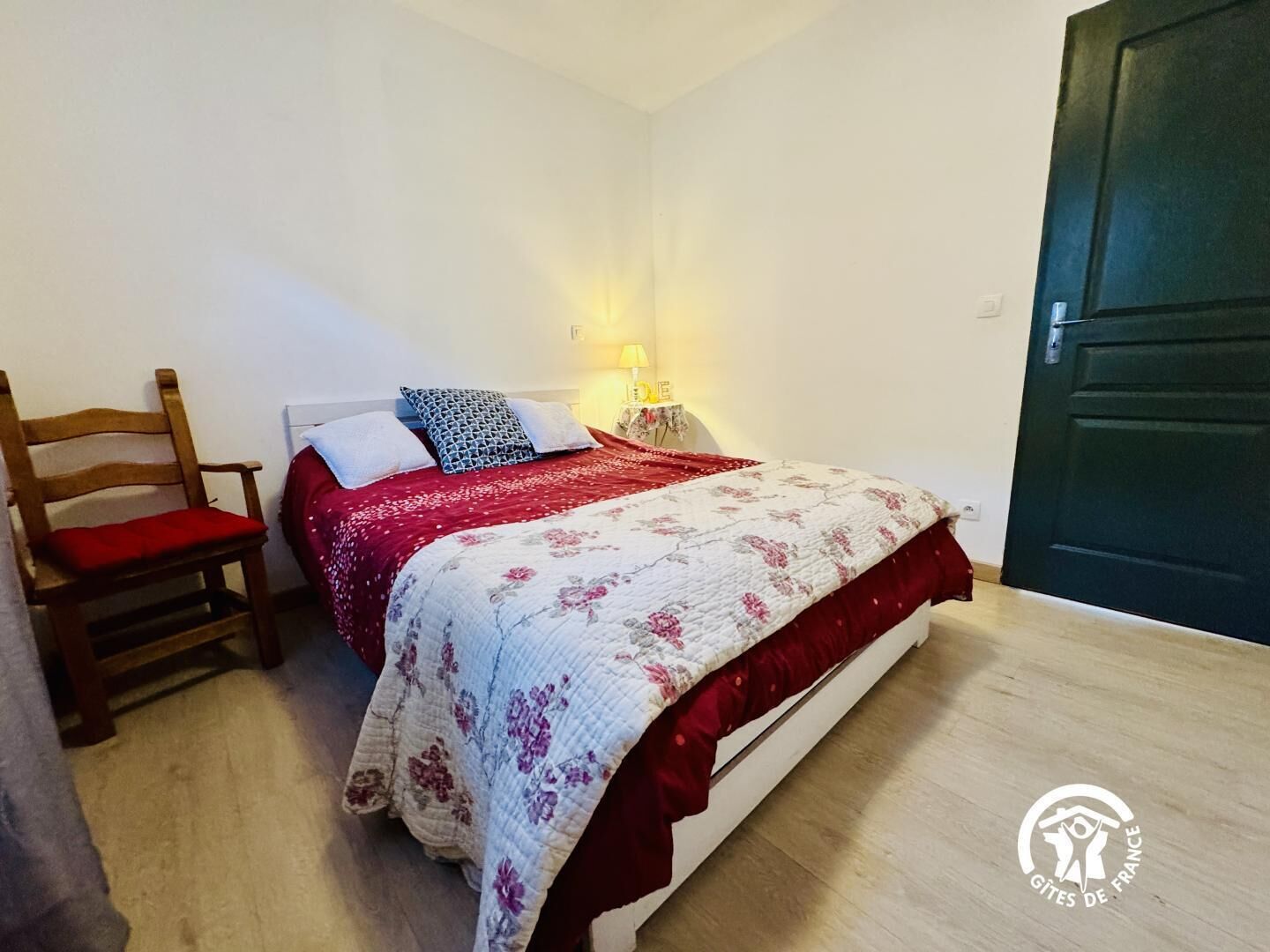 A bedroom with a bed, a chair, a nightstand with a lamp, and a door.