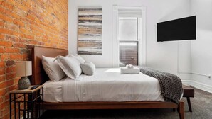 A bedroom with a brick wall, a bed with white bedding, a bedside table with a lamp, and a TV mounted on the wall. - *NEW* Walk 2 Short North | Hot Tub | Easy Parking (Columbus)