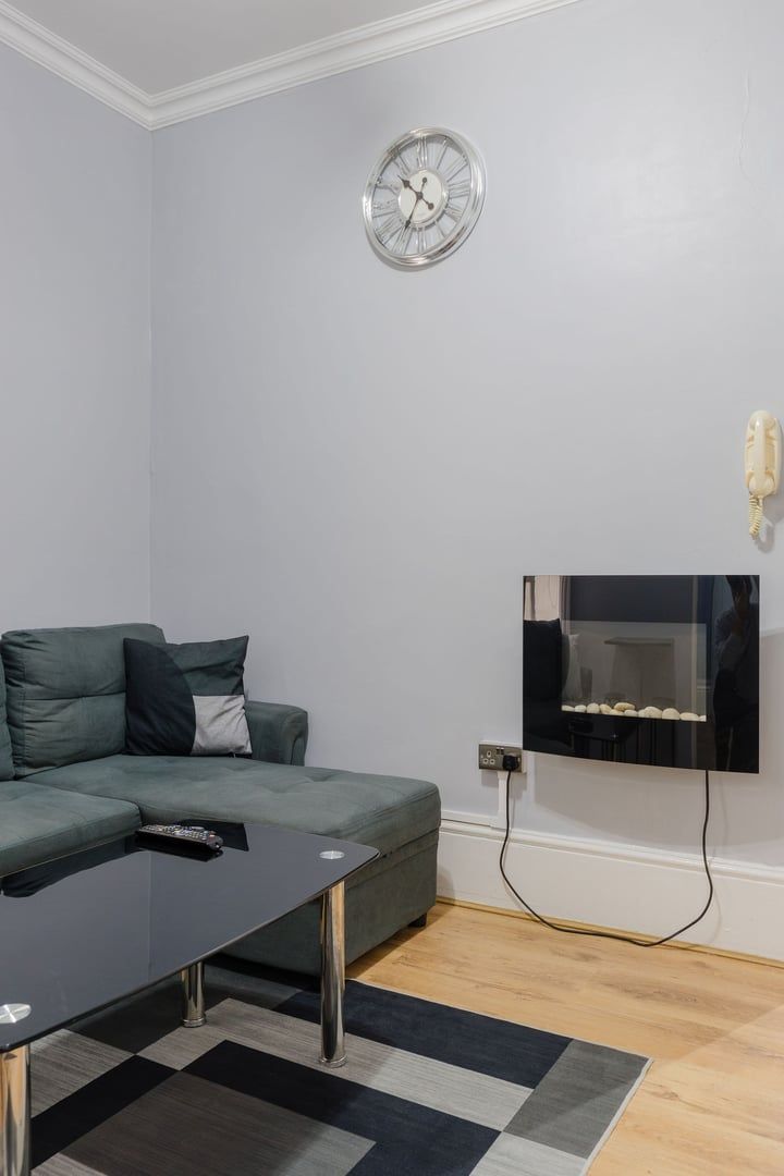 Un salon moderne avec un canapé gris, une table basse noire, un téléviseur fixé au mur et une horloge.