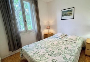 Ein Schlafzimmer mit einem Bett, einem Nachttisch, einer Lampe, einem Fenster mit Vorhängen und einem gerahmten Bild an der Wand.