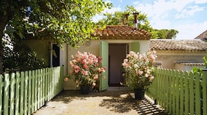 A house with a tiled roof, a green door, and a white picket fence. There are two potted plants with pink flowers in front of the door. - Chai du Canal - SALLELES D'AUDE (SALLELES D AUDE)