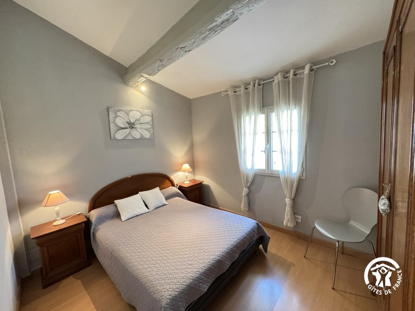 Une chambre à coucher comprenant un lit, deux lampes de chevet, une fenêtre avec des rideaux, une chaise et une œuvre d’art accrochée au mur.