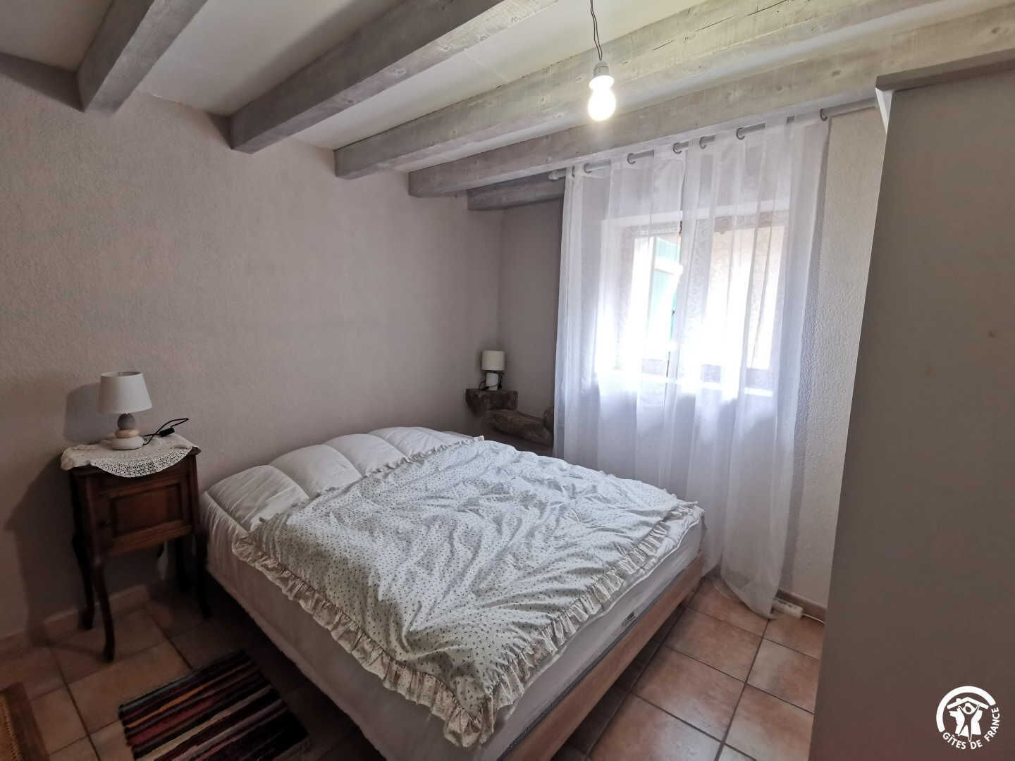 Une chambre à coucher avec un lit, une table de chevet avec une lampe, une fenêtre avec des rideaux et une poutre en bois au plafond.