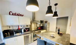 Une cuisine moderne dotée d’appareils électroménagers en acier inoxydable, d’un îlot central, de luminaires suspendus et d’une enseigne « Cuisine » sur le mur.