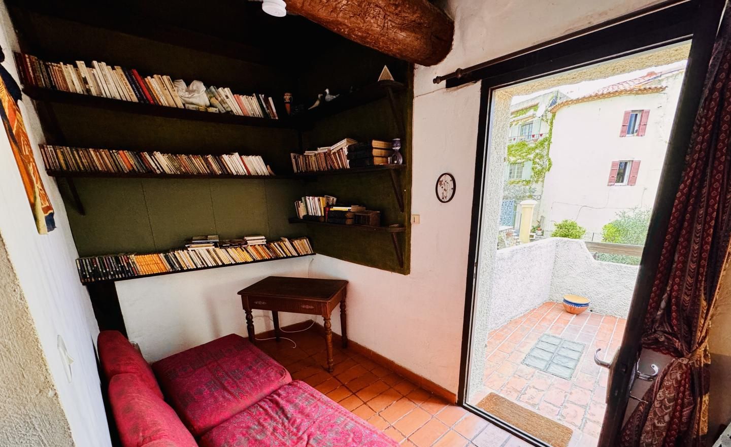 A cozy room with a red sofa, a wooden table, and bookshelves filled with books. A window reveals a view of a building and a courtyard.