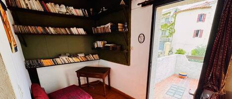 A cozy room with a red sofa, a wooden table, and bookshelves filled with books. A window reveals a view of a building and a courtyard.