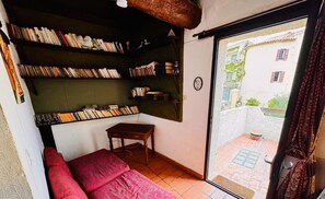 A cozy room with a red sofa, a wooden table, and bookshelves filled with books. A window reveals a view of a building and a courtyard.
