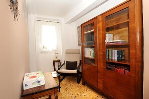 A room with a wooden bookshelf, a chair, a small table with a floral tablecloth, and a window with curtains.