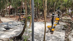 Un espace extérieur aménagé pour la pratique d’exercices physiques, équipé de divers appareils de musculation, et entouré d’arbres et d’un chemin en béton.