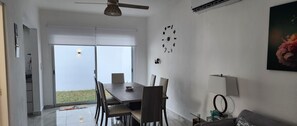 A modern living room with a dining area, a ceiling fan, a wall clock, and a large window offering a view of the outdoors. - A peaceful Yance Villa, 5 min from the beautiful Puerto Morelos beach (Cancún)
