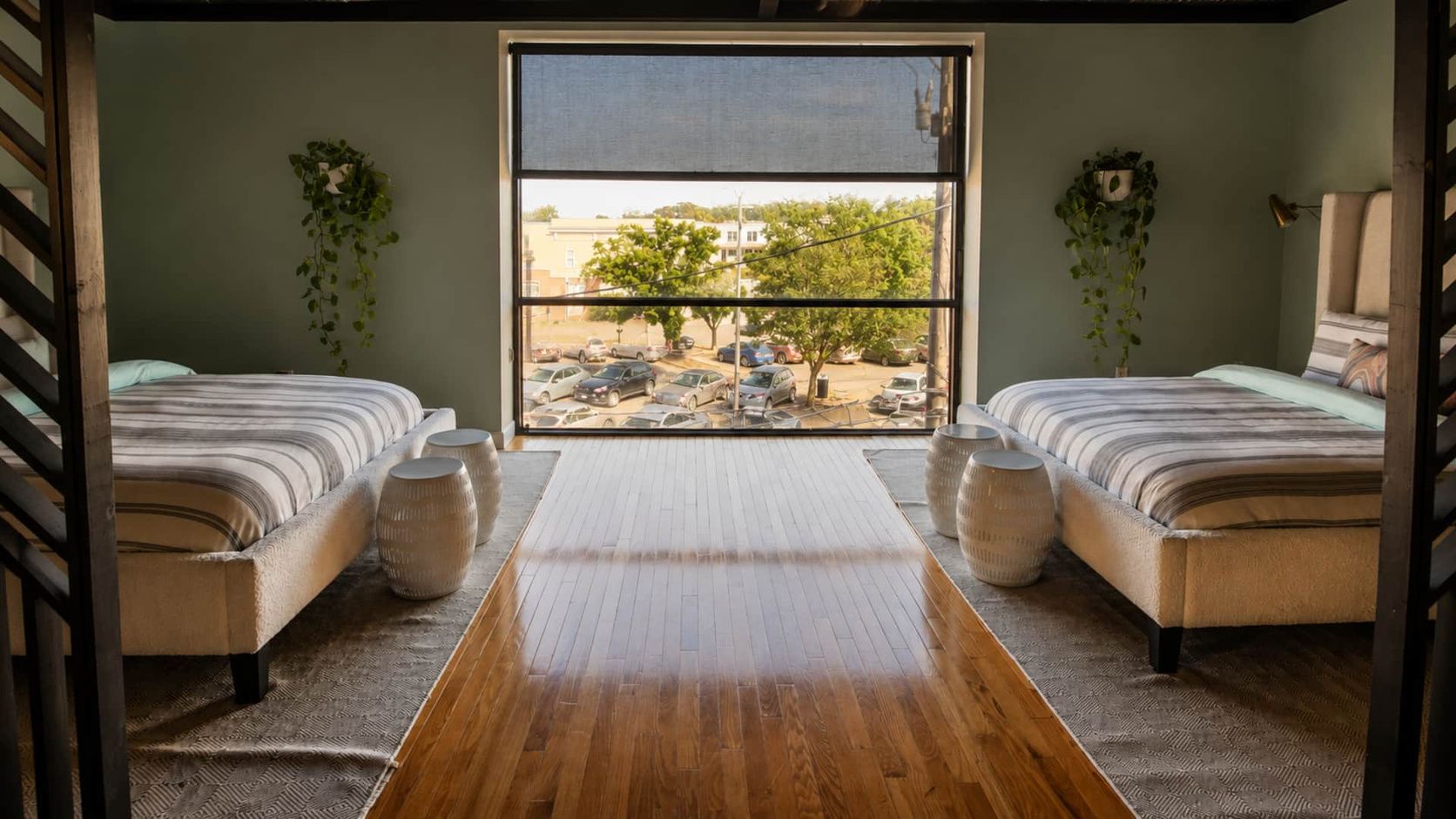 A room with two beds, a large window, and a view of parked cars and trees outside.