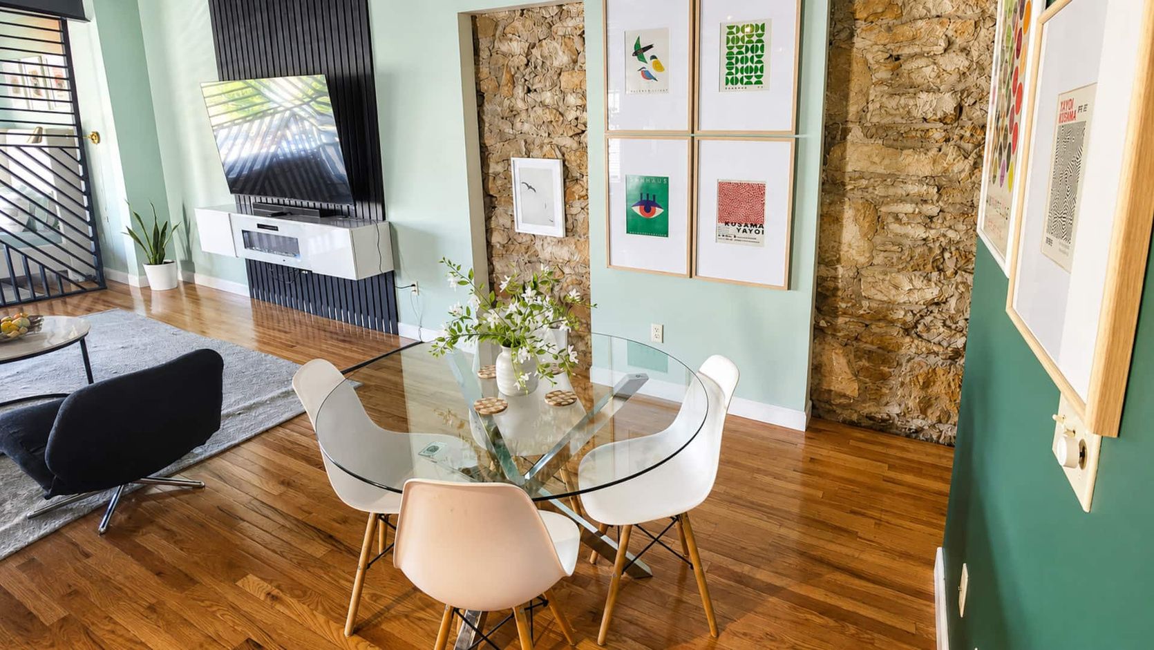 A modern living room with a glass table, two chairs, a flat-screen TV, a stone wall, and framed artwork.