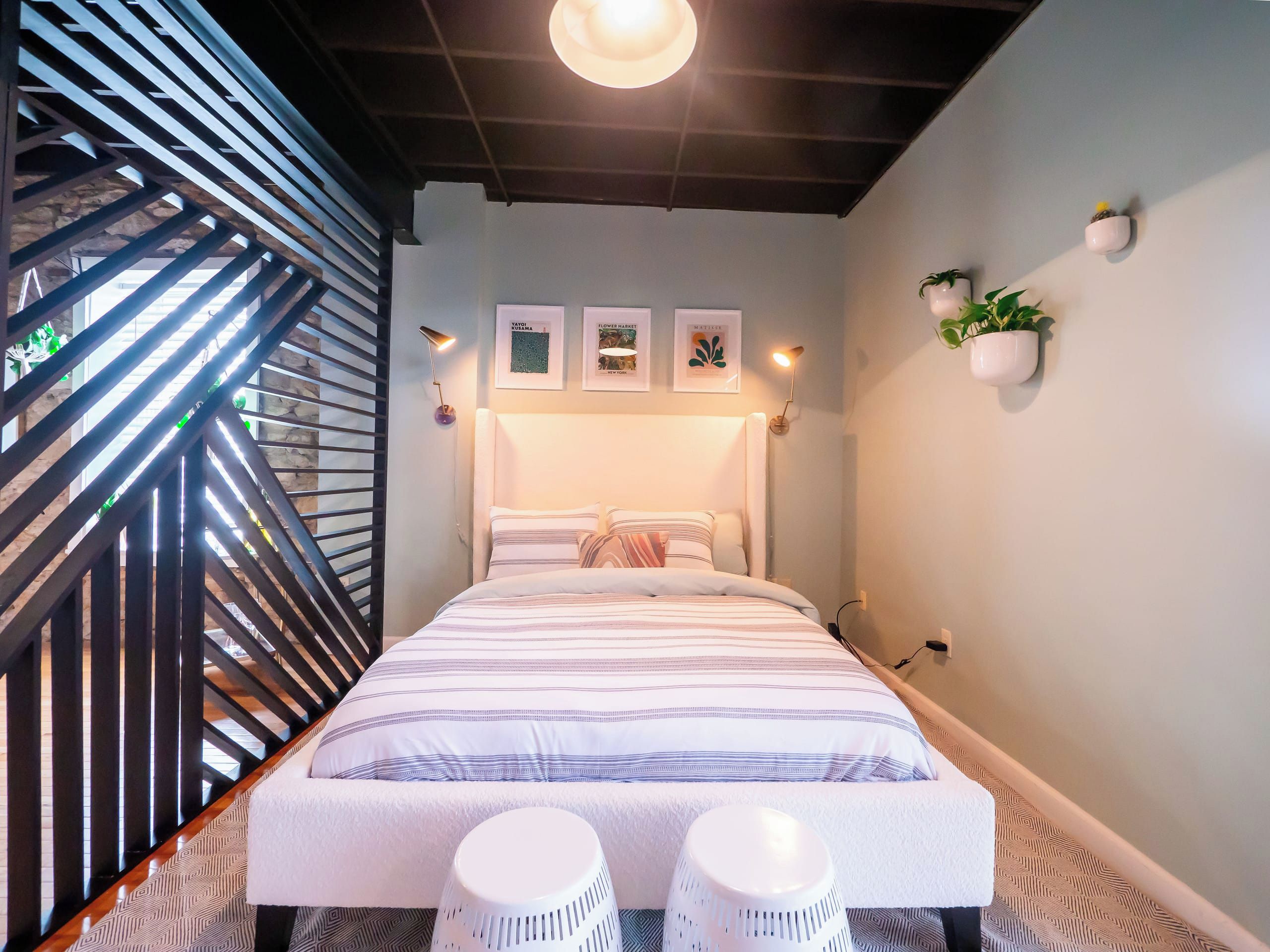 Une chambre moderne avec un lit, deux tabourets blancs, un escalier et des lampes fixées au mur.