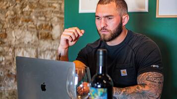 Un homme est assis à une table, devant un ordinateur portable, avec un verre de vin et une bouteille de vin.