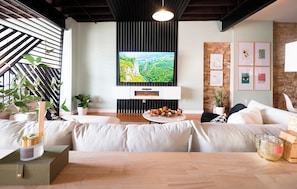 A modern living room with a large flat-screen TV mounted on a wall, a sofa set, a coffee table with decorative items, and a wooden dining table.
