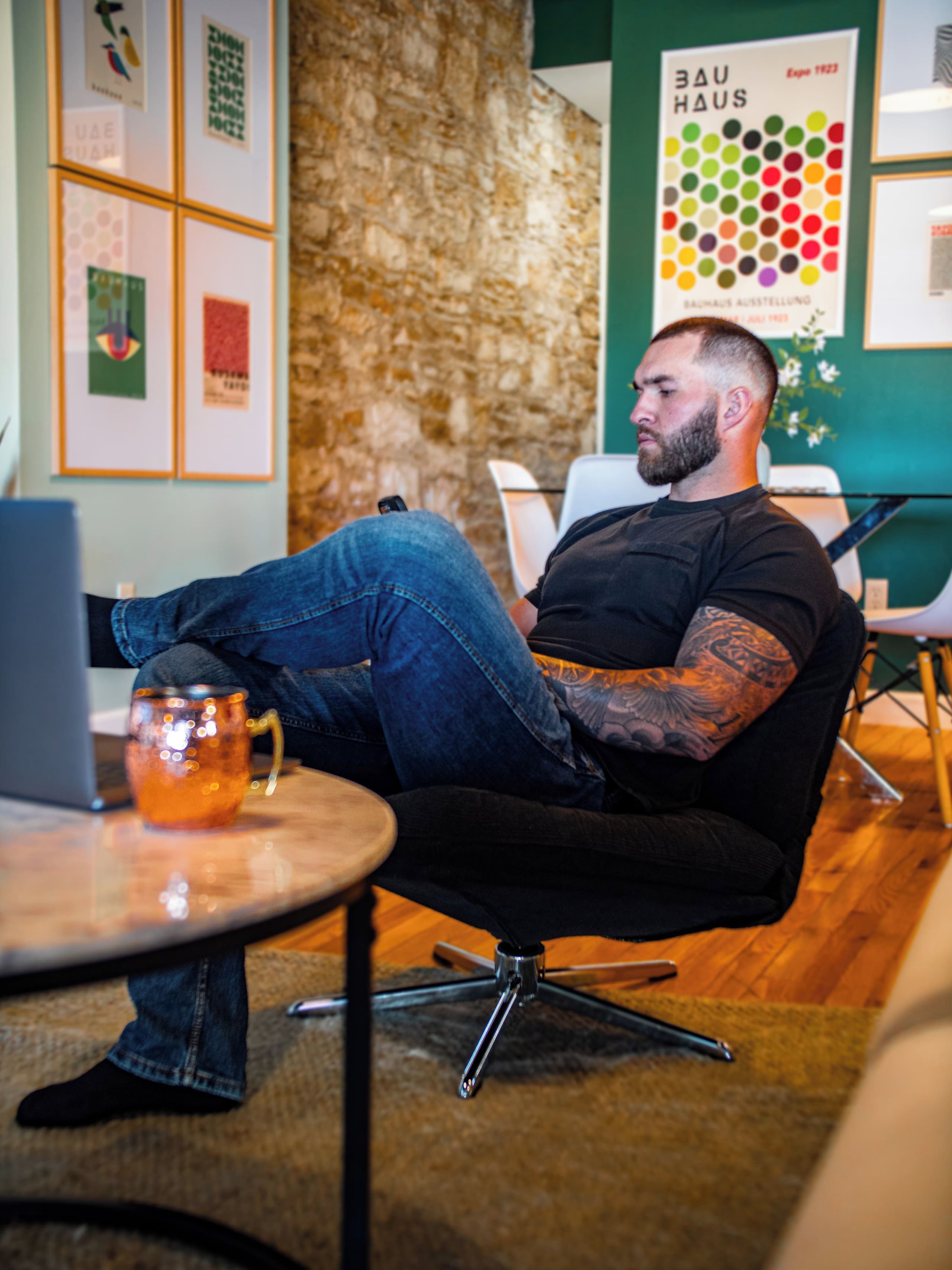 A man seated in a modern chair with a laptop on a table, a mug on the floor, and a poster on the wall.