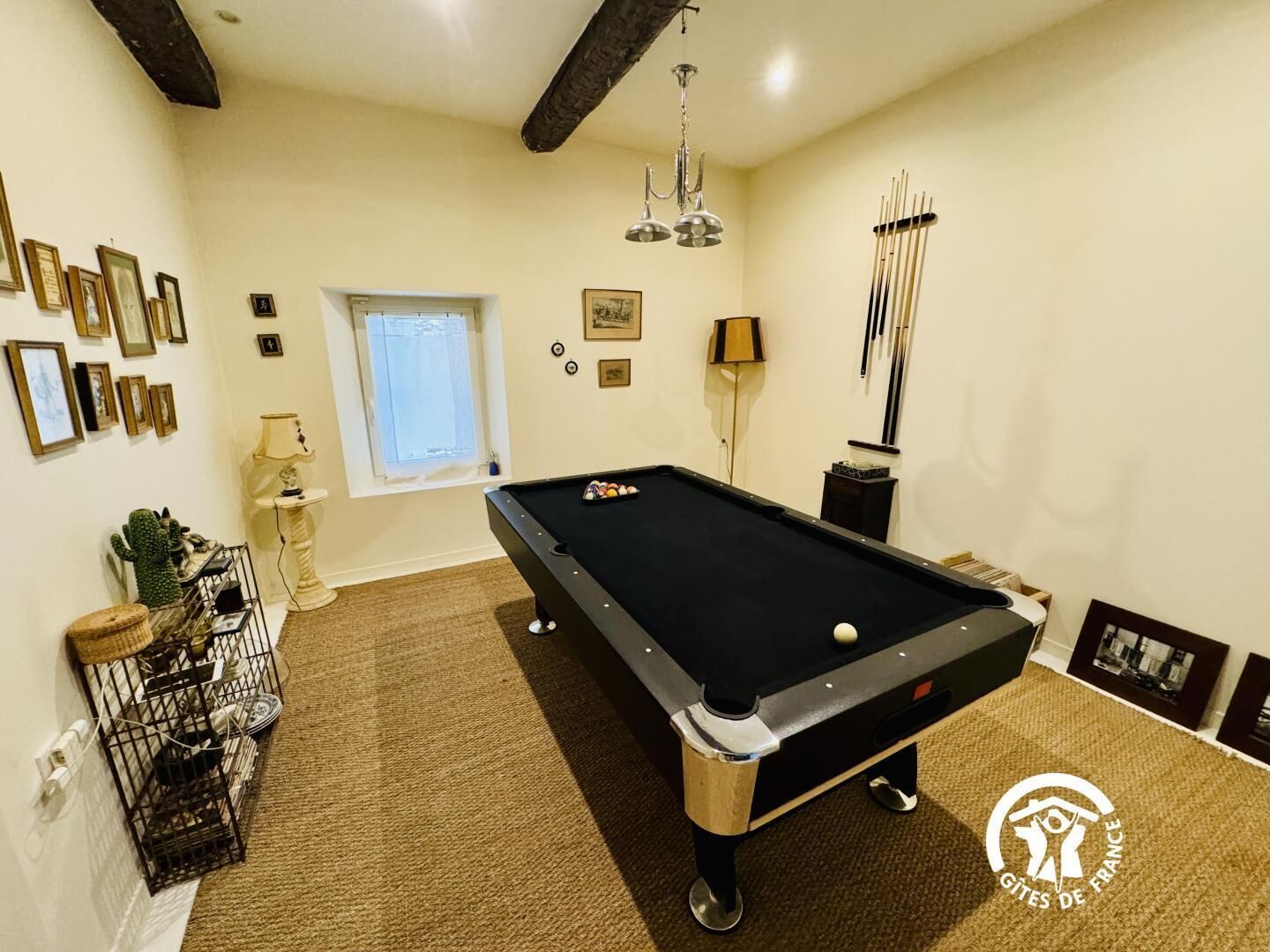 A pool table in a room with framed pictures on the walls, a window, and a lamp.